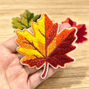 Op de afbeelding: Een esdoornblad-patch met een textuurrijke borduursel in een verloop van geel, oranje en donkerrood met een witte rand. Het ontwerp bevat donkerrood gestikte nerven. Andere blad-patches in groen en rood zijn zichtbaar op de achtergrond.