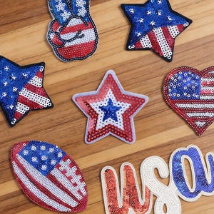 Może przedstawiać: Kolekcja naszywek z cekinami o patriotycznych wzorach, w tym znak pokoju, gwiazdy, serce, piłka do futbolu amerykańskiego oraz napis USA. Każda naszywka prezentuje czerwone, białe i niebieskie kolory w układzie przypominającym flagę USA.