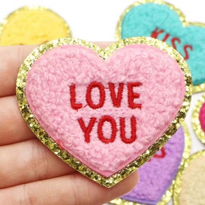 May include: A pink heart-shaped patch with the words "LOVE YOU" embroidered in red. The patch has a gold glitter border and a fuzzy texture. Other heart-shaped patches are visible in the background.