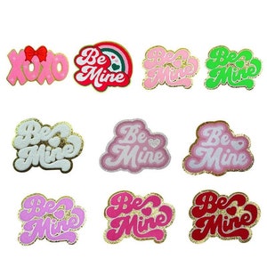 May include: A set of 12 iron-on patches with a Valentine's Day theme. The patches are pink, red, white, and green with gold glitter accents. The patches feature the text "Be Mine" and "XOXO".