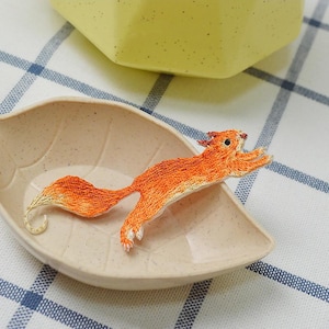 May include: An embroidered orange squirrel with a fluffy tail, set in a leaf-shaped dish. The squirrel is detailed with various shades of orange and white. The text "Longancraft" is above the dish.