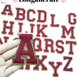 Pode incluir: Uma coleção de patches de letras universitárias bordadas em bordô e dourado. A letra "A" é segurada em uma mão, com outras letras visíveis ao fundo. A marca "Longancraft" está no topo.
