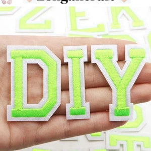 Puede incluir: Letras de fieltro verde que deletrean "DIY" con contornos blancos. Las letras están en una mano.