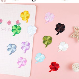 May include: Assortment of ten embroidered iron-on patches in various colors, each featuring a four-leaf clover design. The patches are arranged on a pink surface.