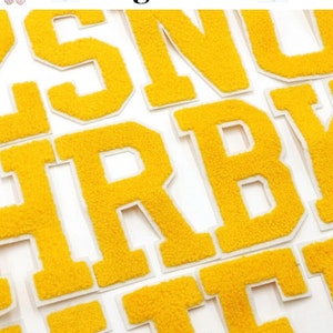 May include: Close-up of yellow chenille letter patches with white borders. The letters are arranged on a white surface. The brand name "Longancraft" is displayed at the top of the image.