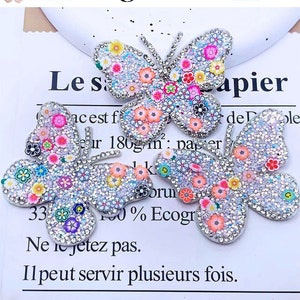 May include: Four butterfly-shaped embellishments with sparkling silver glitter and colorful floral designs. The butterflies are adorned with small, vibrant flowers in shades of pink, yellow, blue, and green. The background is a white paper with French text.