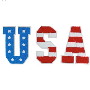 Può includere: USA scritto in grandi lettere di feltro. Le lettere sono blu, rosse e bianche, con un motivo a stelle sulla lettera blu e un motivo a strisce sulle lettere rosse e bianche.