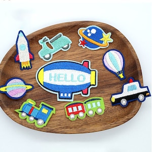 May include: A set of nine colourful embroidered iron-on patches featuring various transportation themes, including a rocket, a scooter, a hot air balloon, a police car, a train, and a blimp that says "HELLO".