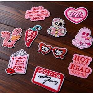 May include: A selection of embroidered patches with book-related themes. The patches display phrases such as "Book Girlie," "Busy Reading Books," and "Hot Girls Read Books." Other designs include a bookworm, a skull reading a book, and heart-shaped glasses.