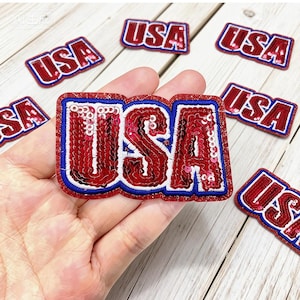 May include: A collection of red, white, and blue sequined "USA" patches. The patches have a red sequined fill, white border, and blue outline. The patches are on a light wood surface. The brand name "Longancraft" is at the top.