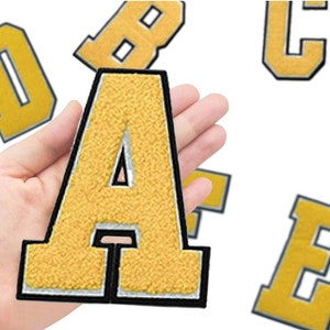 May include: A set of yellow felt iron-on letters, including the letters A, B, C, D, and E. The letters have a black outline and a white inner outline. The letters are shown on a hand.