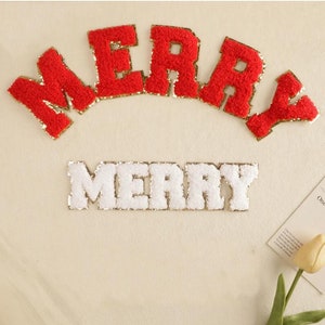 Può includere: Due toppe di feltro rosso e bianco con la scritta "Merry" e bordatura dorata glitterata. Le toppe sono su uno sfondo beige.