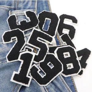 Op de afbeelding: Een set zwarte vilt strijk-op-nummers op een blauwe denim achtergrond. De nummers omvatten 0, 1, 2, 3, 4, 5, 6, 7, 8 en 9. De nummers zijn wit omrand.