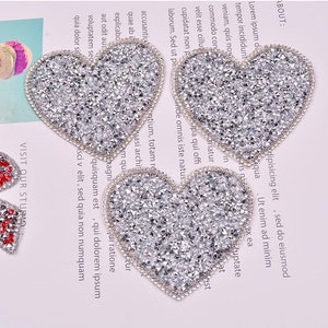 May include: Four silver glitter heart-shaped iron-on patches. The patches are about 3 inches wide and 3 inches tall. The patches are perfect for adding a touch of sparkle to clothing, bags, or other crafts.