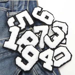 Può includere: Un set di dieci toppe in feltro bianco da stirare a forma di numeri da 0 a 9. I numeri hanno un contorno nero e sono disposti in una pila su uno sfondo di denim blu.