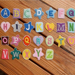 Peut inclure: Écussons brodés de l'alphabet, de formes variées : étoiles, cœurs et cercles. Chaque écusson présente une lettre de l'alphabet dans un design coloré. La marque "Longancraft" est affichée en haut.