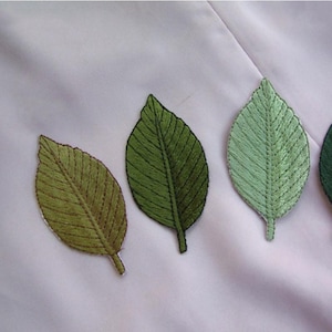 Pode incluir: Aplicações de folhas bordadas em tons de verde, do azeitona ao verde claro, são dispostas sobre um tecido branco. As folhas têm costuras detalhadas, mostrando as veias e as bordas. A marca "Longancraft" está no topo.