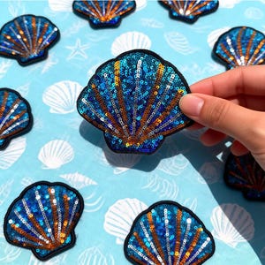 May include: Sequin seashell patches in shades of blue and orange, with a black border. The patches are arranged on a light blue background with seashell patterns. The text "Longancraft" is at the top.