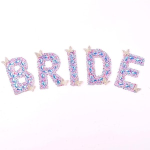 Może przedstawiać: Słowo "BRIDE" zapisane błyszczącymi, klejnotowymi literami. Każda litera jest ozdobiona iryzującymi klejnotami w odcieniach różu, błękitu i fioletu, ze złotymi akcentami motyli. Marka "Longancraft" znajduje się na górze.