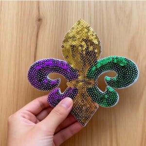 Puede incluir: Un diseño de flor de lis con lentejuelas doradas, verdes y moradas. El diseño se sostiene sobre un fondo de madera. El texto "Longancraft" se muestra encima de la flor de lis.