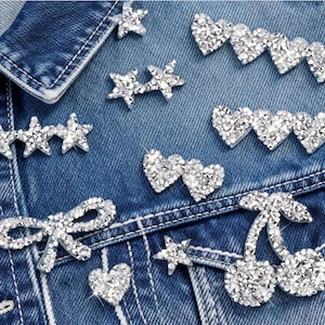 Peut inclure: Gros plan sur une veste en jean ornée de strass étincelants. La veste présente diverses formes, notamment des étoiles, des cœurs, des nœuds et des cerises. Le texte "Longancraft" est affiché en haut. Les strass transparents reflètent la lumière.