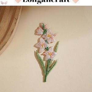 May include: Embroidered floral patch featuring a cluster of pink and yellow flowers with green leaves and stem. The patch has the text "Longancraft" above the flowers. The background is a solid, light beige color.