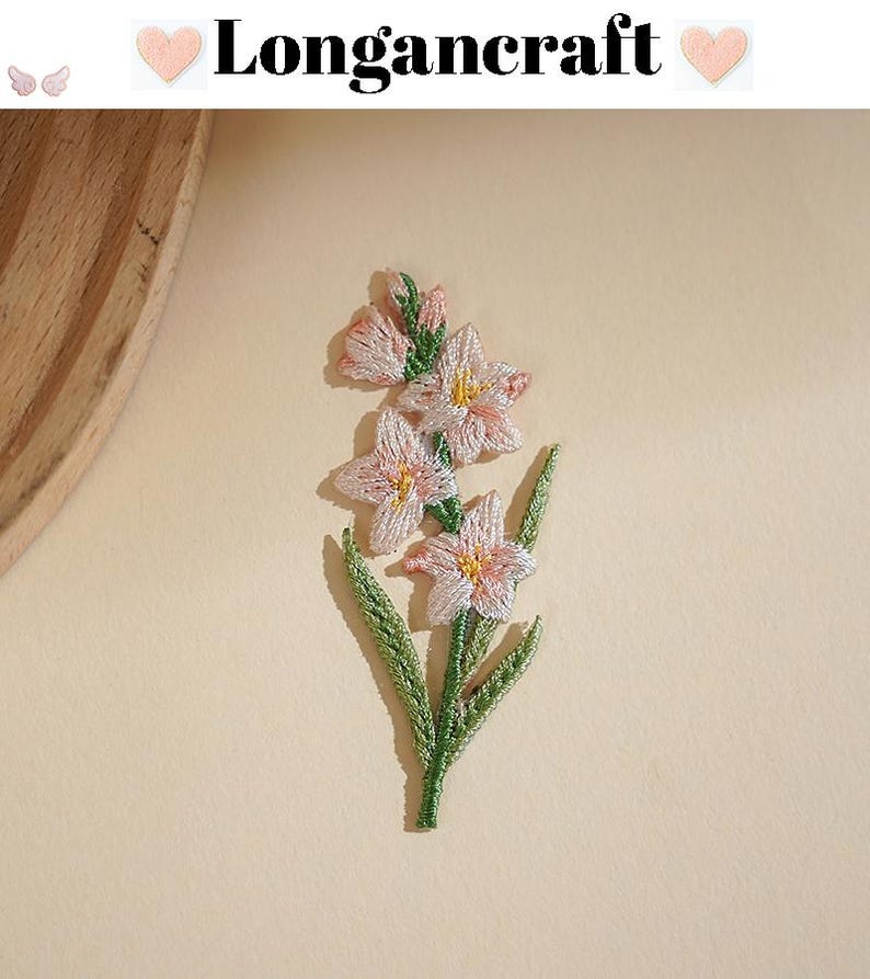 May include: Embroidered floral patch featuring a cluster of pink and yellow flowers with green leaves and stem. The patch has the text "Longancraft" above the flowers. The background is a solid, light beige color.