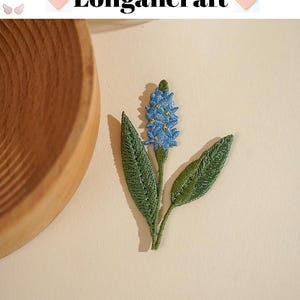 May include: Embroidered floral patch featuring a blue flower with yellow accents and green leaves. The patch has the text "Longancraft" at the top. The patch is on a light beige background.