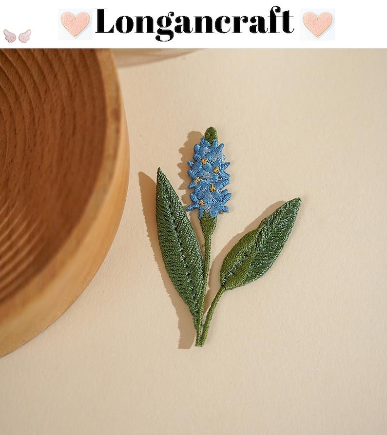 May include: Embroidered floral patch featuring a blue flower with yellow accents and green leaves. The patch has the text "Longancraft" at the top. The patch is on a light beige background.