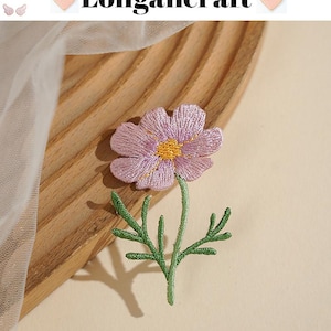 May include: Embroidered purple flower with a yellow center and green stem and leaves. The flower is on a wooden surface. The text "Longancraft" is above the flower.