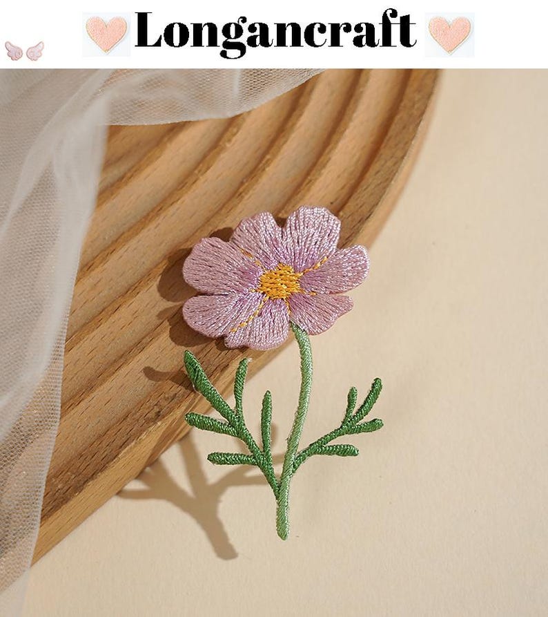 May include: Embroidered purple flower with a yellow center and green stem and leaves. The flower is on a wooden surface. The text "Longancraft" is above the flower.
