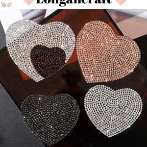 May include: Four heart-shaped patches covered in rhinestones. The patches are in black, white, rose gold, and clear. The patches are on a brown surface.