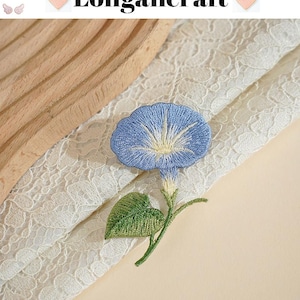 May include: Embroidered blue morning glory flower with green leaves and stem. The flower is set against a backdrop of white lace and a light beige surface. The text "Longancraft" is at the top.