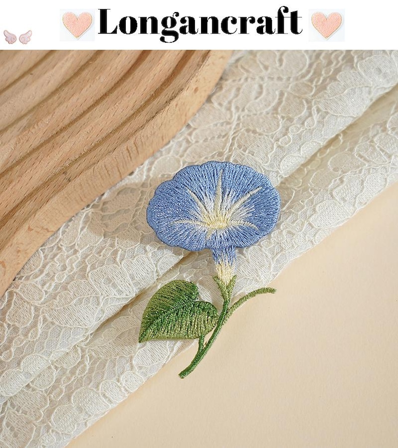 May include: Embroidered blue morning glory flower with green leaves and stem. The flower is set against a backdrop of white lace and a light beige surface. The text "Longancraft" is at the top.