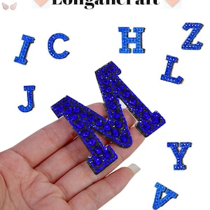 Pode incluir: Letras do alfabeto de strass azul, incluindo as letras I, C, J, M, H, Z, L, Y e V. As letras estão sobre um fundo branco.