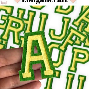 Pode incluir: Um close-up de uma letra A de feltro verde com contorno de feltro amarelo. A letra está sendo segurada em uma mão. A letra é uma das muitas letras na imagem, todas as quais são verdes com contornos amarelos.
