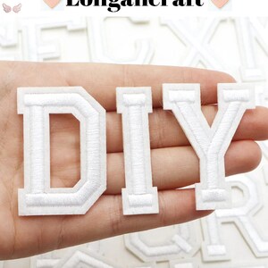 May include: White felt letters spelling out "DIY" in a collegiate style font. The letters are thick and have a white outline. The letters are held in a hand.