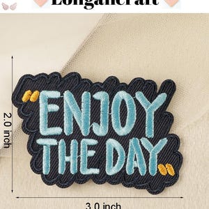 Pode incluir: Um remendo de ferro bordado azul e preto com o texto "Enjoy the Day" em um contorno branco. O remendo mede 7,6 cm de largura e 5,1 cm de altura.
