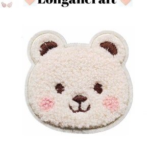 May include: White fuzzy bear face applique with brown embroidered eyes, nose, and mouth. The bear has pink cheeks.
