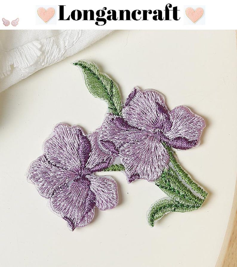 May include: Embroidered floral patches featuring two purple flowers with green leaves. The patches have a detailed stitch design and are set against a white background. The text "Longancraft" is at the top.
