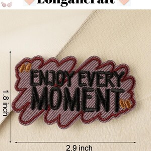 Pode incluir: Etiqueta bordada roxa com o texto "Enjoy Every Moment" em letras pretas com detalhes dourados. A etiqueta mede 2,9 polegadas de largura e 1,8 polegadas de altura.