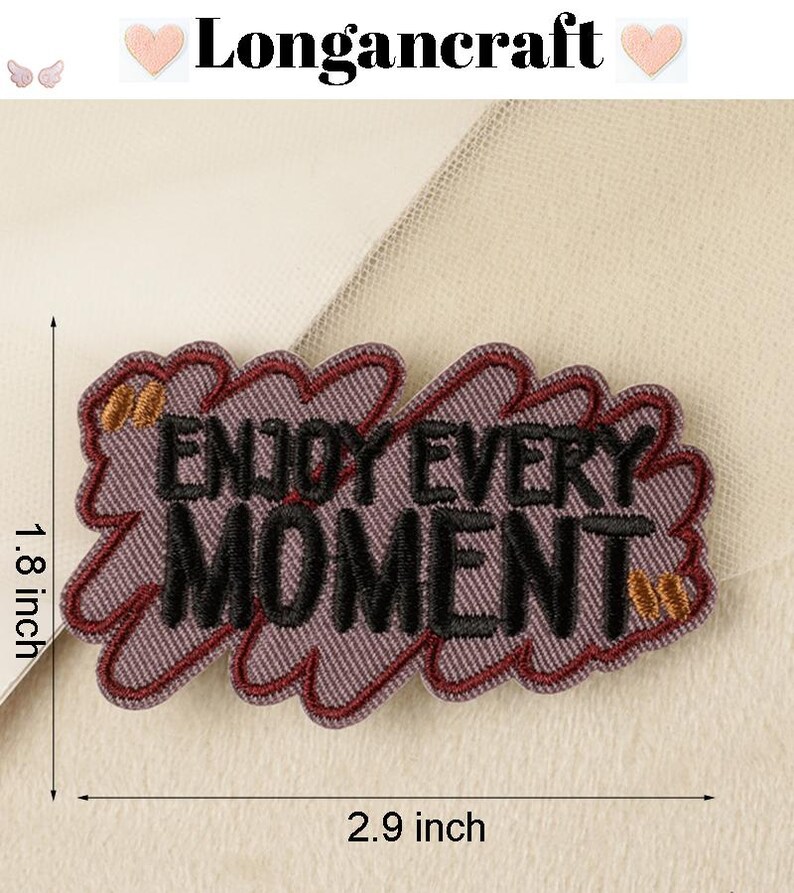 Pode incluir: Etiqueta bordada roxa com o texto "Enjoy Every Moment" em letras pretas com detalhes dourados. A etiqueta mede 2,9 polegadas de largura e 1,8 polegadas de altura.