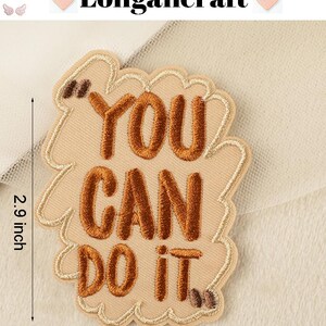 Pode incluir: Etiqueta bordada de ferro bege com o texto "You Can Do It" em fio marrom. A etiqueta mede 5,6 cm de largura e 7,4 cm de altura.