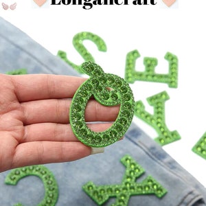 May include: Green iron-on letters with rhinestones. The letters are C, O, S, E, Y, and X. The letters are on a denim background.