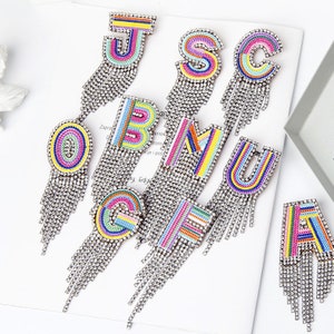 Può includere: Orecchini a forma di lettere dell'alfabeto. Ogni orecchino presenta una lettera diversa, impreziosita da ricami colorati e strass scintillanti. Lunghe frange di strass pendenti aggiungono movimento e stile a questi orecchini.