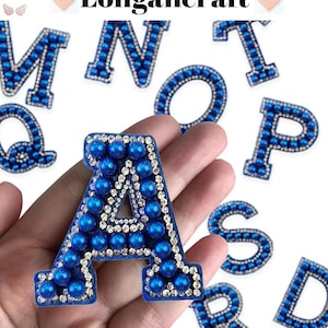 Pode incluir: Um conjunto de patches de letras do alfabeto azul e branco. As letras são feitas de contas azuis e strass branco. As letras estão dispostas em uma ordem aleatória em uma mão.