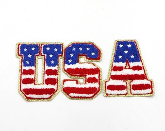 May include: USA spelled out in large, red, white, and blue letters with gold glitter trim. Each letter is decorated with a pattern of the American flag.