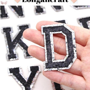 May include: Black and silver glitter iron-on letters in the style of varsity letter patches. The letters are in uppercase and include the letters N, K, Y, and D. The letters are on a white background.