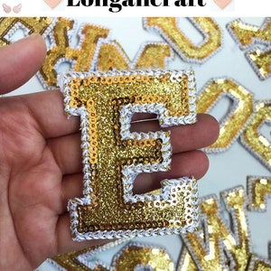May include: Gold glitter letter E with white trim. The letter is made of sequins and is surrounded by other gold glitter letters.