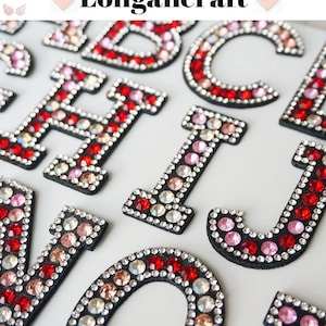 Op de afbeelding: Een set strijkbare alfabetletters bedekt met rode, roze en heldere strasssteentjes. De letters zijn in hoofdletters en zijn in willekeurige volgorde gerangschikt. De letters bevinden zich op een witte achtergrond.